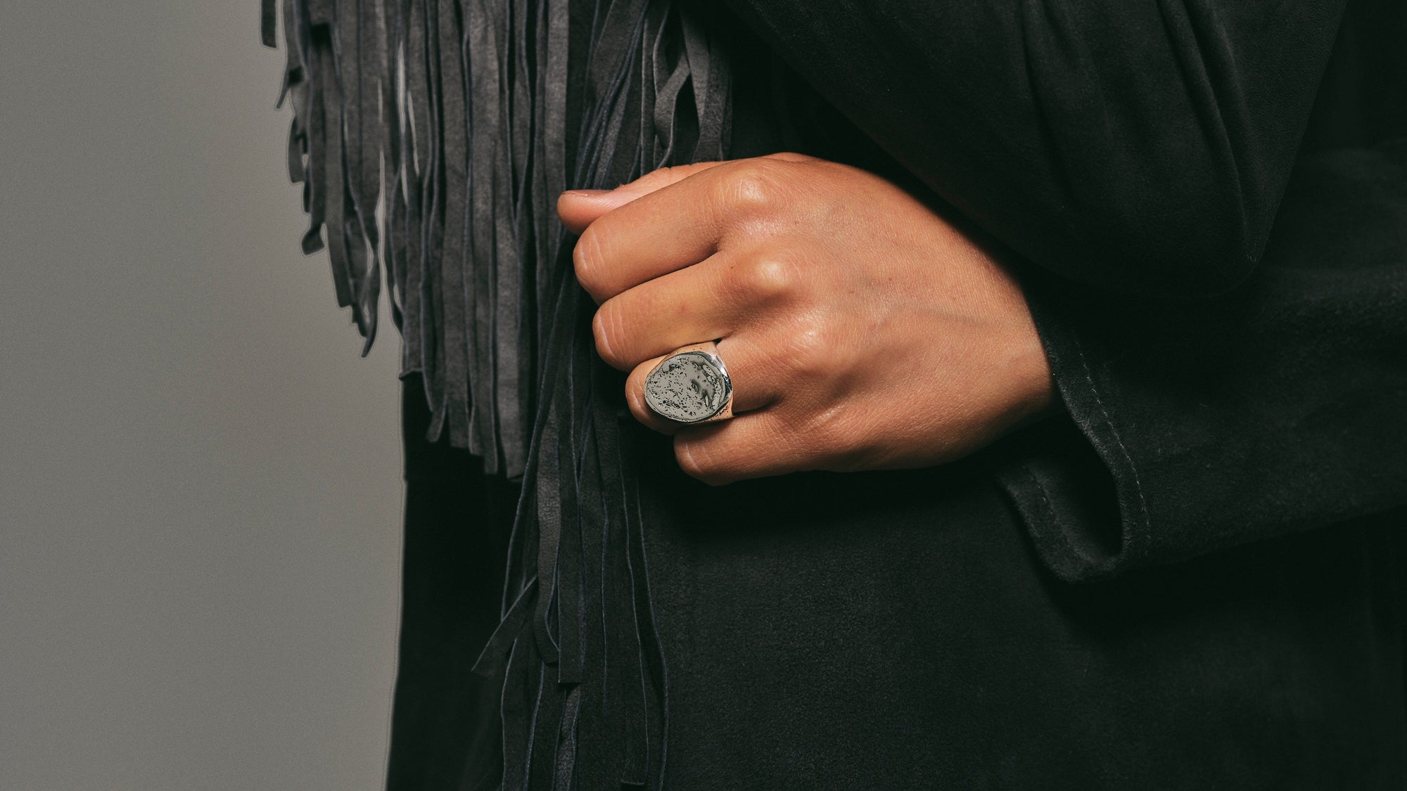 Person wearing a black coat with fringe details and a ring on a gray background